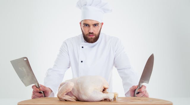 Portrait-of-a-serious-male-chef-cook-standing-with-knifes-and-chicken-on-the-table-isolated-on-a-white-background