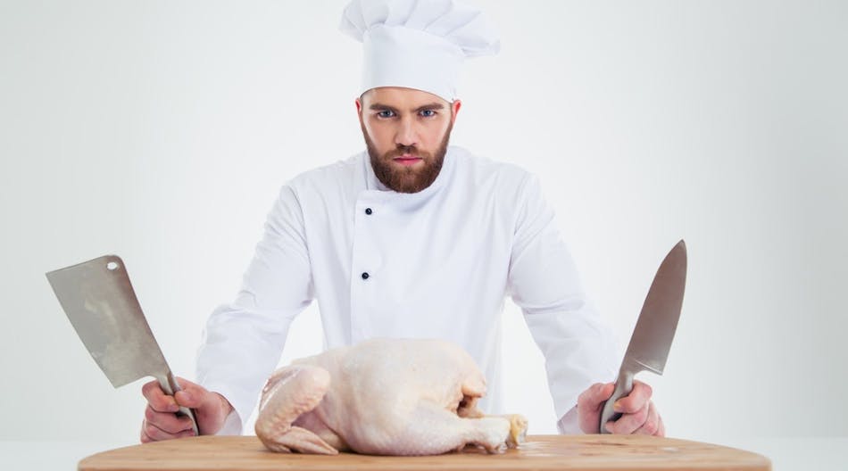 Portrait-of-a-serious-male-chef-cook-standing-with-knifes-and-chicken-on-the-table-isolated-on-a-white-background