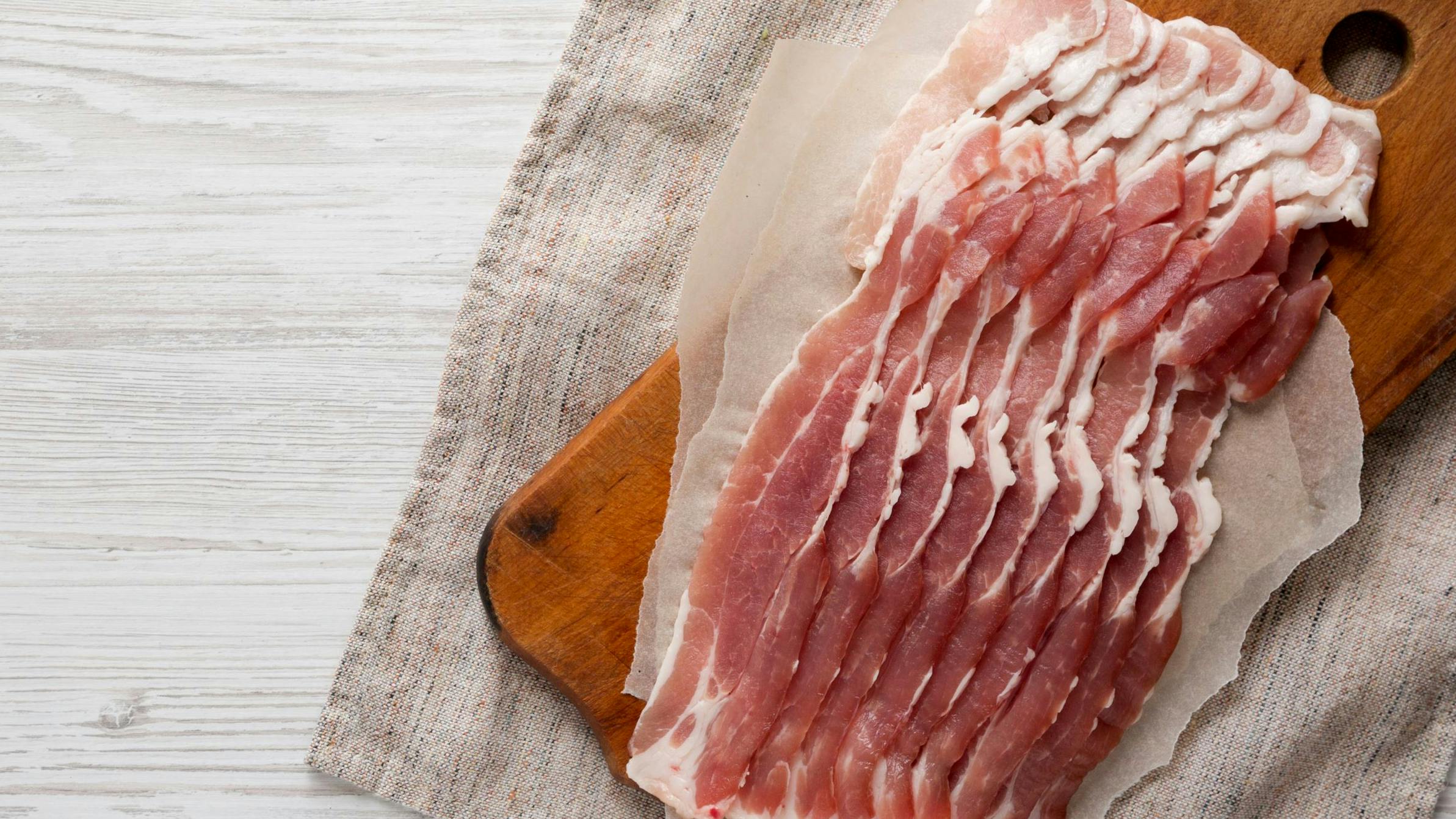 Uncooked Raw Bacon on a rustic wooden board on a white wooden background, top view.