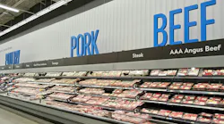 Grocery store meat department with long refrigerated display cases filled with packaged meat products. Large blue text on the white wall above reads 'POULTRY,' 'PORK,' and 'BEEF' from left to right. Grocery store meat department with long refrigerated display cases filled with packaged meat products. Large blue text on the white wall above reads 'POULTRY,' 'PORK,' and 'BEEF' from left to right.