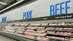 Grocery store meat department with long refrigerated display cases filled with packaged meat products. Large blue text on the white wall above reads 'POULTRY,' 'PORK,' and 'BEEF' from left to right. Grocery store meat department with long refrigerated display cases filled with packaged meat products. Large blue text on the white wall above reads 'POULTRY,' 'PORK,' and 'BEEF' from left to right.
